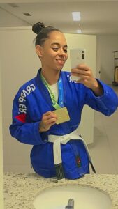 Na foto, Tamires está vestindo um kimono azul com a palavra "GRAPPLER" escrita nas mangas. Ela está segurando uma medalha de ouro olhando para o espelho com o celular na mao.